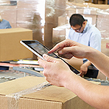hands holding tablet in warehouse with stacks of boxes