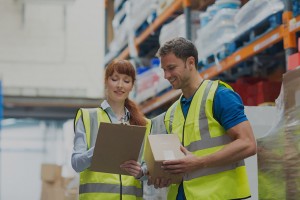 woman and man in warehouse in reflective vests holding clipboard and cardboard box
